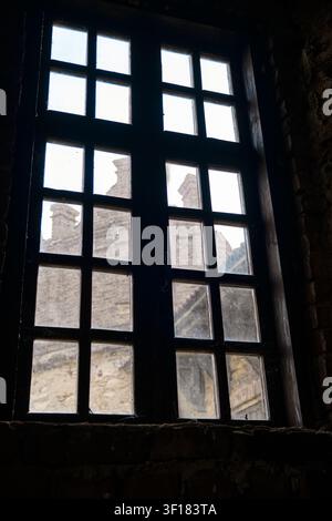Eine niedrige Innenansicht eines alten Holzfensters mit mehreren Scheiben, das auf ein rustikales Steingebäude hinausblickt und Schatten in einen dunklen Ziegelraum wirft. Stockfoto