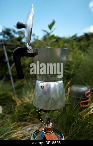 Ein klassischer Aluminium-Moka-Topf-Geysir-Kaffeebereiter mit offenem Deckel steht auf einem tragbaren Gasbrennerkocher im Freien im grünen Gras während eines Berges Stockfoto
