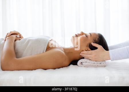 Junge schwarze Frau entspannt sich auf dem Spatisch, während der Therapeut ihren Kopf massiert Stockfoto
