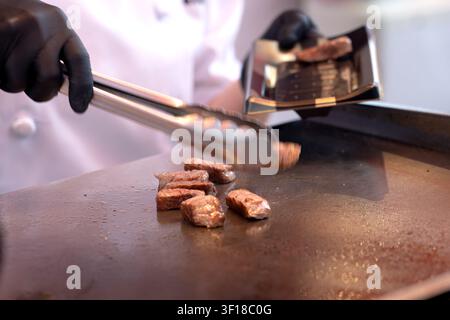 Ein professioneller Koch mit schwarzen Handschuhen verwendet Metallzangen, um saftige, gewürfelte Steakhappen auf einer heißen Teppanyaki-Grillplatte zuzubereiten Stockfoto