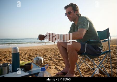 Mann sitzt in einem Klappstuhl auf einem Sandstrand und macht Kaffee über einem tragbaren Campingkocher, umgeben von Thermoskannen und Tassen, und genießt einen entspannten Ozeanmorgen Stockfoto