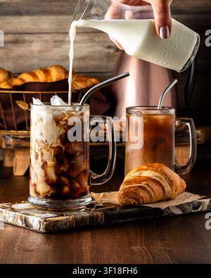 Milch in ein Glas Eiskaffee gießen, serviert mit Croissants. Stockfoto