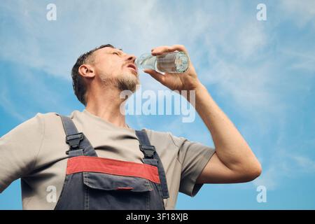 Mann in Latzhose hydriert sich während der Arbeit im Freien unter heißem Sommerhimmel Stockfoto