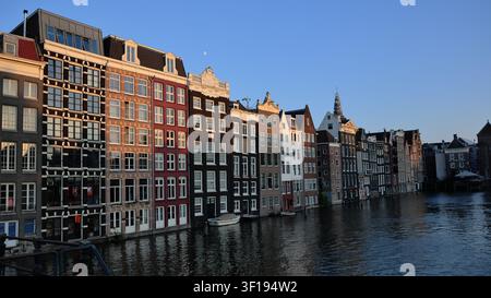 Amsterdamer Kanalhäuser bilden eine lange Uferreihe am Damrak, ihre schmalen Fassaden und unterschiedlichen Dachlinien spiegeln sich im dunklen Wasser unter einem klaren blauen Himmel wider. Stockfoto