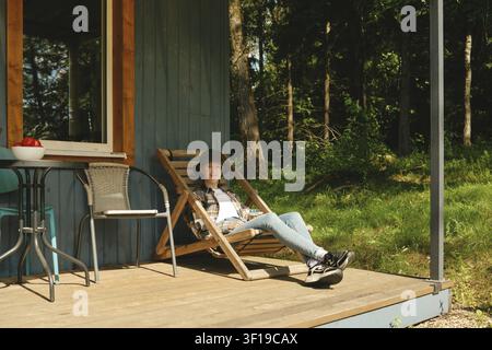 Fröhliche junge Frau entspannt sich auf einem Liegestuhl vor einer Holzhütte im Wald. Ein kleiner Tisch mit Obst steht in der Nähe Stockfoto