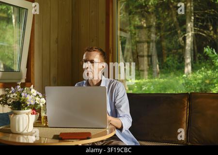 Mittealter Mann sitzt an Tisch und nutzt Laptop in Holzhütte und konzentriert sich auf Online-Lernen. Sonnenlicht kommt durch Fenster und wirft Licht auf Holz Stockfoto