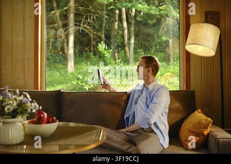 Mittelalter Mann sitzt auf braunem Sofa in Holzhütte. Er liest ein E-Book in einem klimatisierten Raum, während draußen die Mittagshitze herrscht. Sonnenlicht dringt durch d Stockfoto