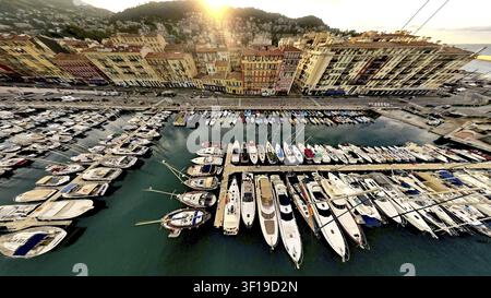 Ein malerischer Hafen bei Sonnenuntergang mit Reihen von Booten und farbenfrohen Gebäuden vor einer hügeligen Kulisse, Drohnenfotografie, Nizza, Frankreich Stockfoto