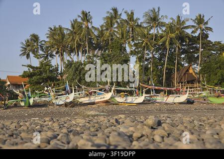 Traditionelle, farbenfrohe balinesische Fischerboote namens Jukung reihen sich an einem felsigen Kieselstrand mit hohen Palmen und einer ruhigen Lagune auf. An den Ufern der Stockfoto