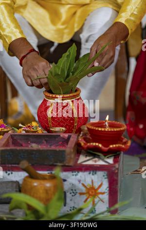 Eine Person führt eine hinduistische religiöse Zeremonie durch und legt Blätter in einen geschmückten Kalash-Topf neben einer brennenden Diya-Öllampe Stockfoto