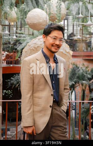Eleganter asiatischer Mann in stilvollem beigem Blazer und blauem Hemd, posiert in grünem Café im Innenraum, professionelles Porträt eines gutaussehenden Gentlemans mit Brille Stockfoto