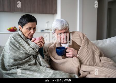 Trauriger älterer Mann, Frau, die an einer Grippe leidet, sitzt auf der Couch, bedeckt mit Decken, trinkt heißen Tee Stockfoto