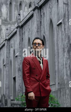 Attraktiver asiatischer Mann in rotem Zweireiher-Anzug, der an einer historischen grauen Steinkathedrale vorbeigeht, klassischer Heritage-Gentleman-Straßenmode-Stil-Porträt Stockfoto