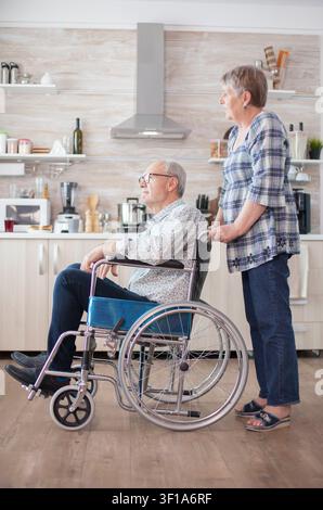Behinderter Senior sitzt im Rollstuhl in der Küche, während seine Frau in der Nähe hilft und damit Unterstützung für Menschen mit Behinderung und eine fürsorgliche Ehe im Alter zeigt. Stockfoto