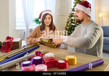 Lächelndes Paar in Weihnachtsmannmützen, das zu Hause gemeinsam ein Weihnachtsgeschenk einpackt Stockfoto