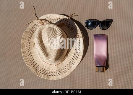 Ansicht von oben für Sonnenschutzutensilien mit einem Strohhut, einer schwarzen Sonnenbrille und einer leeren Kosmetiktube auf einem beigen Hintergrund. Stockfoto