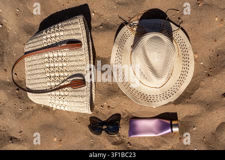 Oberansicht eines Sommer-Strand-Essentials-Layouts mit einem Strohhut, einem geflochtenen Tote, einer Sonnenbrille und einer Kosmetiktube auf einem Sandstrand. Stockfoto