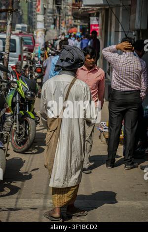 Dhaka, Bangladesch – 27. Februar 2017: Eine Person, die eine hellgraue lange Tunika und einen gelb karierten Lungi trägt, ist von hinten zu sehen, wie sie auf einer überfüllten Str Stockfoto