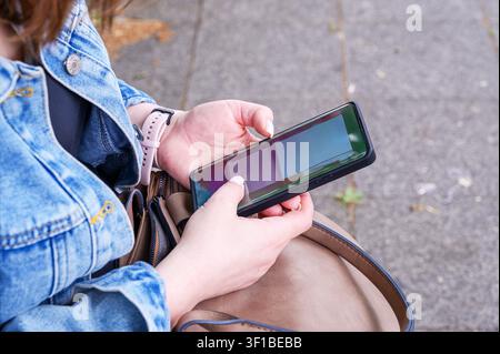 Junge Frau in Jeansjacke, die mit beiden Händen ein Smartphone hält, während sie sitzt. Stockfoto