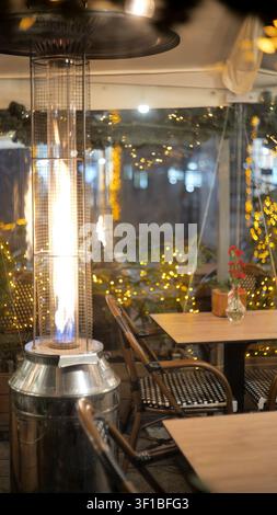 Hoher Gas-Patio-Heizstrahler mit sichtbarer Flamme neben leeren Restauranttischen und geflochtenen Stühlen in einem geschlossenen Außengarten bei Nacht, warme Lichterkette bok Stockfoto