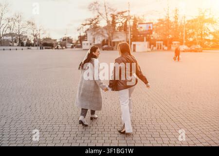 Frauenfreundschaften: zwei junge Frauen halten sich an den Händen und gehen durch einen sonnigen Stadtplatz zur Goldenen Stunde mit Kopierraum Stockfoto