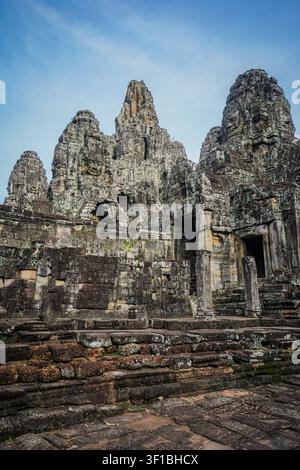 Der antike Bayon-Tempel verfügt über mehrere mehrstufige graue Steintürme mit geschnitzten Gesichtern, die auf einem Sockel aus dunklen, verwitterten Steinblöcken und rotem stehen Stockfoto