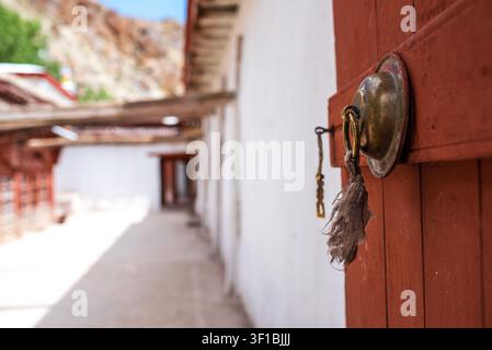 Nahaufnahme eines goldenen Metallklopfers mit einem ausgefransten Tuch, befestigt an einem roten Holzfensterladen. Der Hintergrund zeigt einen verschwommenen Außenkorridor mit weißen Wänden Stockfoto