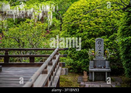 Ein malerischer Blick auf ein traditionelles steinernes Gedenkmal, das mit frischen Blumen geschmückt ist und sich neben einem hölzernen Verandabalcony in einem üppigen grünen Tempelga befindet Stockfoto