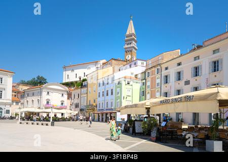 Piran, Slowenien. 26. Mai 2026. Ein sonniger Tartini-Platz ist von farbenfrohen Gebäuden und einem markanten Glockenturm gesäumt. Menschen sind zu sehen, die spazieren gehen und unter Sonnenschirmen im Freien speisen. Stockfoto