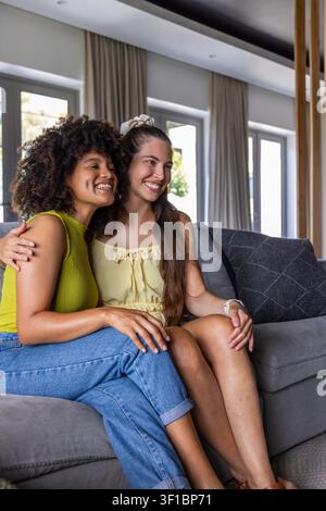 Verschiedene Freundinnen, die eng beieinander auf einem grauen Sofa in einem modernen Wohnzimmer sitzen, bequeme Oberteile tragend. Freundschaft, Zuneigung, Tageslicht, Zuhause, Innenräume, informell, gemütlich Stockfoto