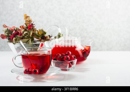 Morgendlicher Herbst-Teetzeit mit rotem Kräutertee in einer durchsichtigen Teekanne, Tasse mit Hagedorn, Hagebutte, getrockneten Blättern im Sonnenstrahl in sanftem Licht in einem weißen Interieur. Stockfoto