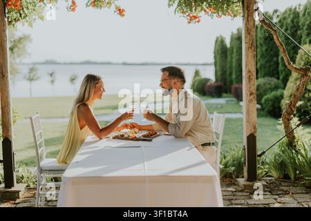 Ein Paar teilt einen freudigen Moment und stößt mit Gläsern an, während es an einem elegant gedeckten Tisch am ruhigen Seeufer sitzt, umgeben von Natur. Stockfoto