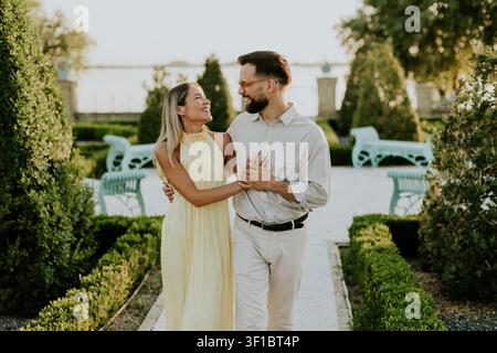 Ein Liebespaar schlendert Hand in Hand durch einen bezaubernden Garten und genießt den warmen Glanz der untergehenden Sonne. Ihr Lächeln spiegelt Freude und Verbundenheit wider. Stockfoto