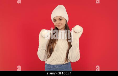 Lächelndes Teenager-Mädchen in warmer Wintermütze und Fäustlingen im Studio. teenager-Wintermädchen isoliert auf Rot Stockfoto
