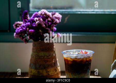 Erfrischender Eiskaffee serviert in einer Café-Atmosphäre mit einem schönen Strauß violetter Statice-Blumen am Fenster Stockfoto