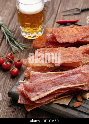 Scheiben geräucherten getrockneten Hühner-, Truthahn- und Schweinefleischs auf Holzbrett mit einem Glas Bier, Rosmarin und Kirschtomaten Stockfoto