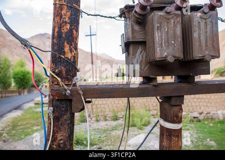 Nahaufnahme eines alten rostigen elektrischen Transformators, der auf rauen Holzpfählen montiert ist. Freiliegende und farbenfrohe Stromkabel sind unsicher befestigt. In der Stockfoto