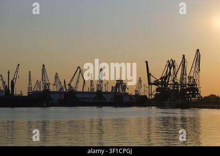 Industriehafenkrane bilden dramatische Silhouetten vor den warmen Tönen der untergehenden Sonne, spiegeln sich im ruhigen Wasser und zeigen globale Logistik Stockfoto