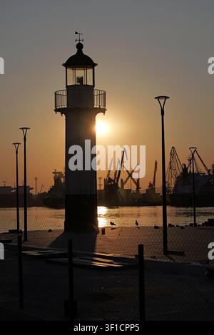 Leuchtturm steht an einem Pier bei Sonnenuntergang, wirft warmes Licht auf das Wasser, während Hafenkrane und Schiffe Silhouetten im Hintergrund bilden, signif Stockfoto