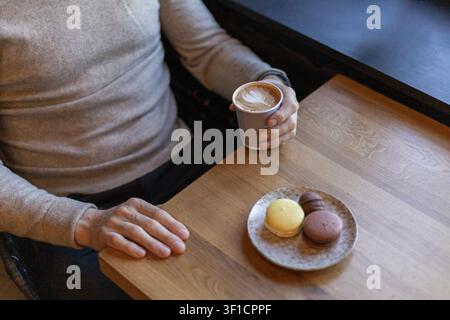 Scharfer, hochwinkliger Schuss eines Mannes in einem grauen Pullover, der an einem Holztisch sitzt und eine Papptasse mit Latte-Art-Kaffee neben einem Teller mit Col hält Stockfoto