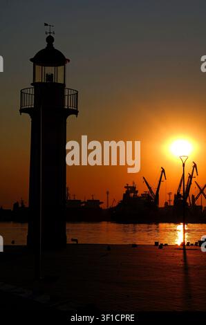 Leuchtturm, der als Silhouette vor einem lebhaften orangefarbenen Sonnenuntergangshimmel steht, mit dem industriellen Hafen im Hintergrund, der Schiffe und Kräne zeigt, die sich spiegeln Stockfoto