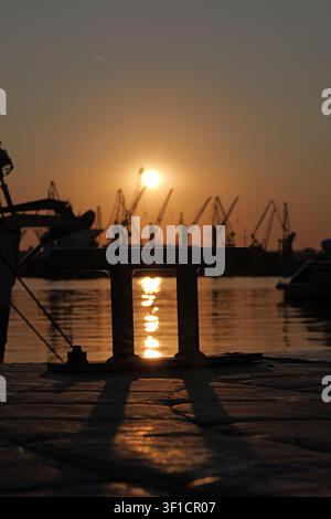 Bollard, der eine starke Silhouette auf dem Pier bildet, mit der lebendigen Sonne, die sich auf dem Wasser spiegelt, zeigt einen geschäftigen Industriehafen mit Ladekränen in t Stockfoto