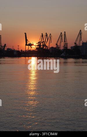 Industrielle Hafenkrane stehen als Silhouette vor einem lebhaften orangefarbenen und gelben Sonnenuntergangshimmel, der sich auf ruhigem Wasser spiegelt, und betonen Themen der globalen t Stockfoto