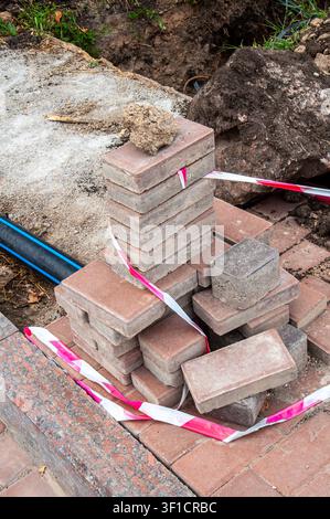 Ein Stapel roter und grauer ineinandergreifender Pflastersteine liegt auf einer Baustelle, die mit rotem und weißem Warnband abgesperrt ist Stockfoto