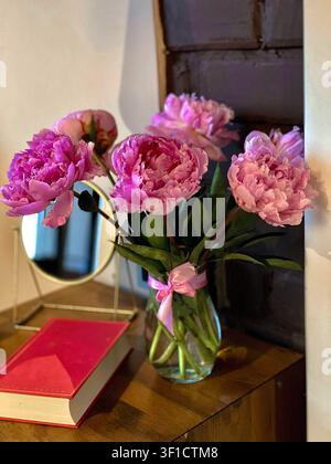 Frische rosa Pfingstrosen in einer Glasvase auf einem Holztisch mit gemütlicher Wohnraumgestaltung. Stockfoto
