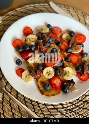 Hausgemachte Mini-Pfannkuchen mit frischen Beeren, Banane und Schokoladenaufstrich auf weißem Teller. Stockfoto