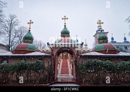 Schneeflocken fallen sanft über ein orthodoxes Kloster und bedecken seine Kuppeln und goldenen Kreuze mit weißem Schnee, während festliche Lichter in der Dämmerung funkeln. Stockfoto