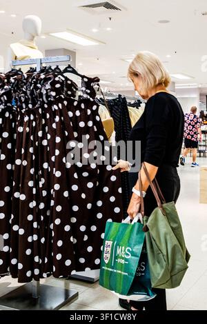 Epsom, London UK, 12. Juli 2026, eine mittelalte Frau hält Einkaufstüten, während sie eine Ausstellung von leichten, gemusterten Sommerkleidern durchstöbert Stockfoto