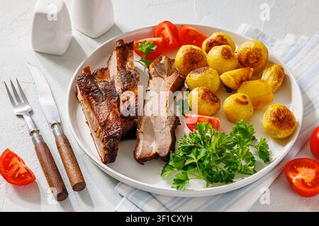 Bayrisch gebackene Schweinerippchen mit ganz gerösteten Kartoffeln, Tomaten, Petersilie auf einem Teller auf weißem strukturiertem Tisch mit Besteck und gelbem Senf,  ver Stockfoto