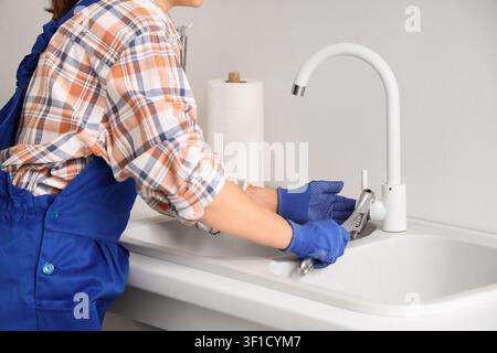 Klempner in blauen Latzhosen und kariertem Hemd repariert einen weißen Küchenwasserhahn mit einem Schraubenschlüssel über einem Waschbecken, der Arbeiter trägt blaue Handschuhe zum Schutz, während pe Stockfoto